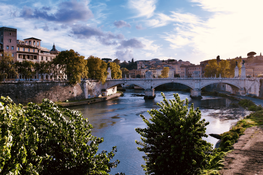 Scenic view of a river with a bridge in the background, framed by trees and historic buildings under a partly cloudy sky.