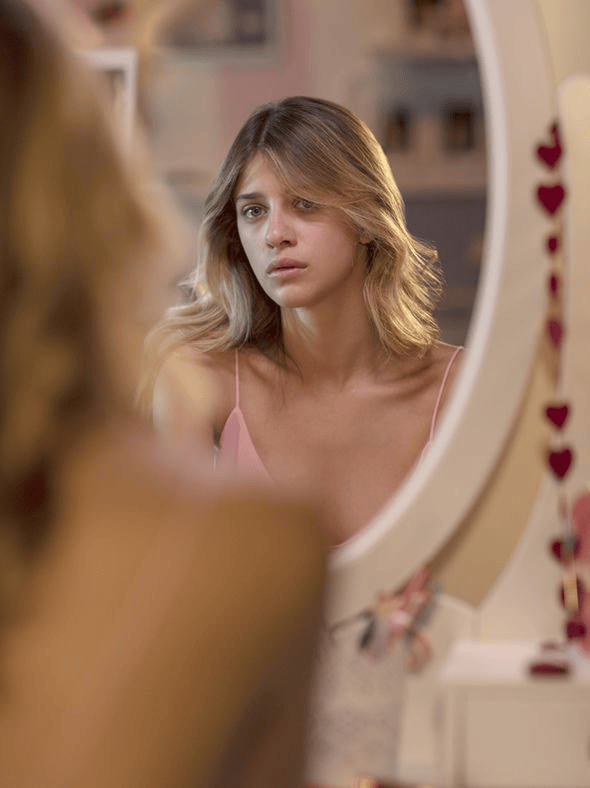 A young woman gazes thoughtfully into a mirror, showcasing a contemplative expression and soft lighting.