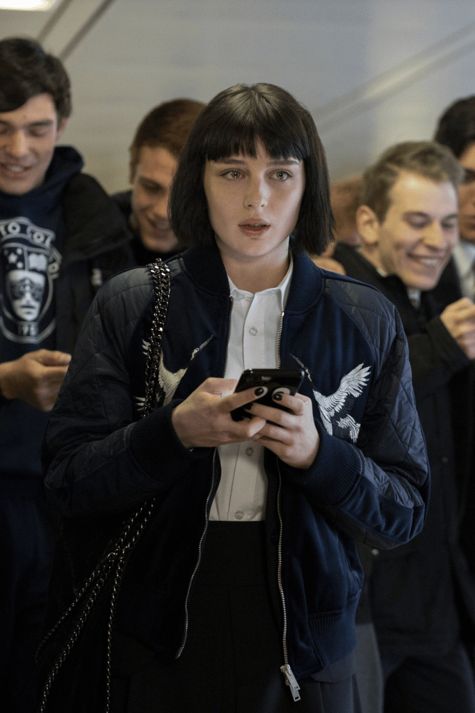 A young woman with short black hair wearing a blue bomber jacket and a white shirt is standing in a crowd, looking at her phone with a neutral expression.