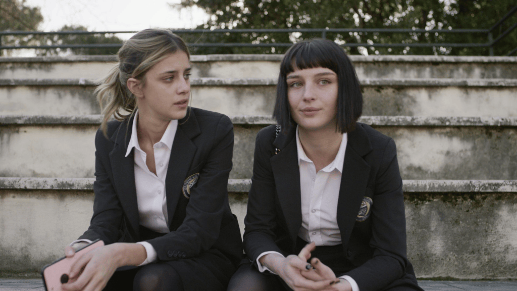 Two teenage girls in school uniforms are sitting on stairs, engaged in conversation. The girl on the left has long wavy hair and is looking thoughtfully to the side, while the girl on the right has straight black hair and is facing the camera with a slight smile.